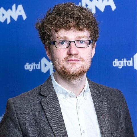 Portrait of Adam McBride, who has curly red-brown hair, glasses and a short beard, wearing a grey blazer and white shirt